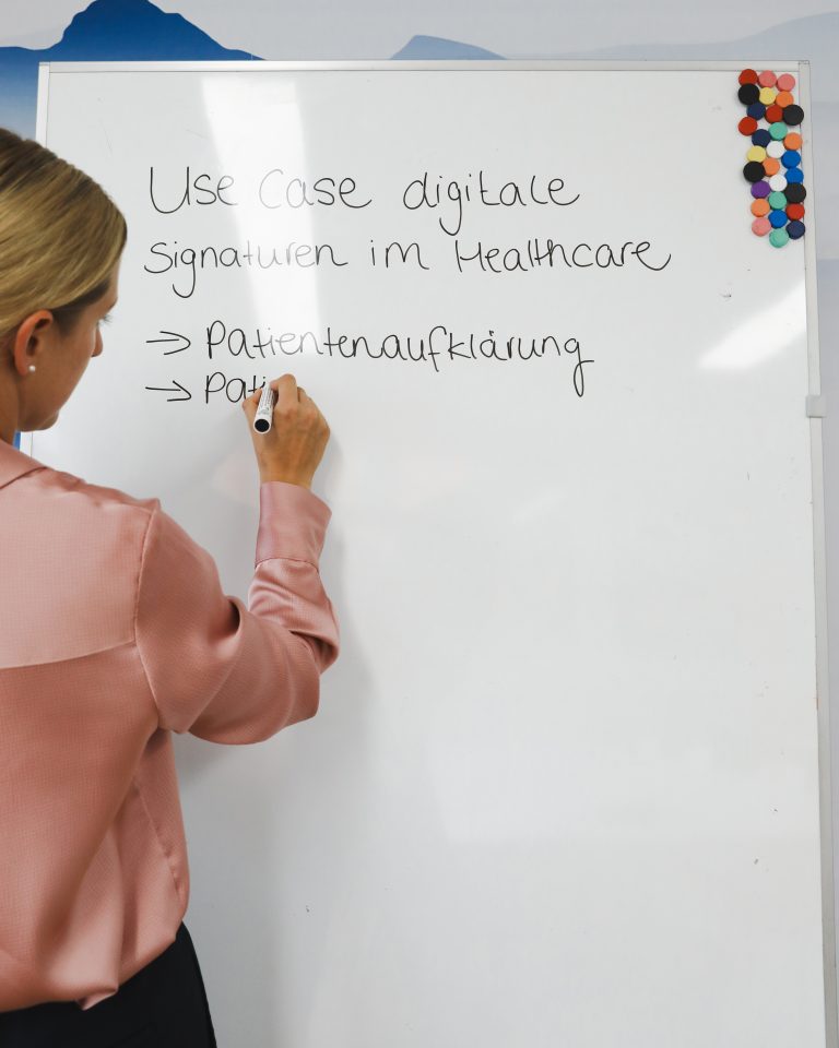 Blonde Frau mit rosafarbener Bluse schreibt auf Whiteboard