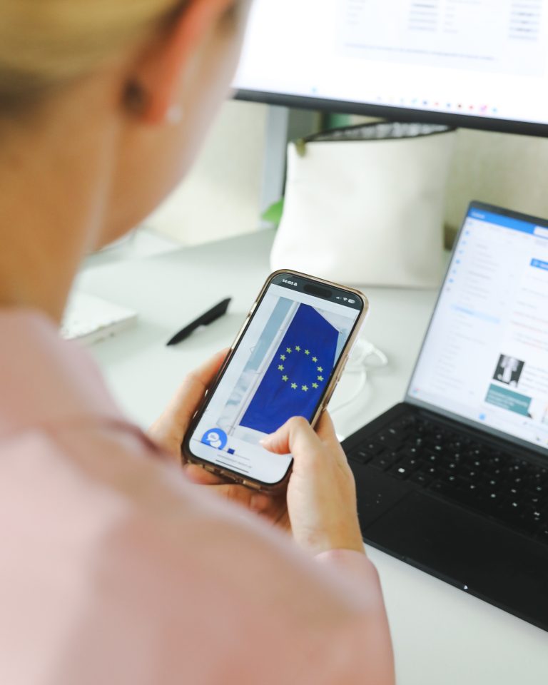 Blonde Frau mit rosafarbener Bluse hält ein Smartphone in der Hand mit der EU-Flagge, digital unterschreiben
