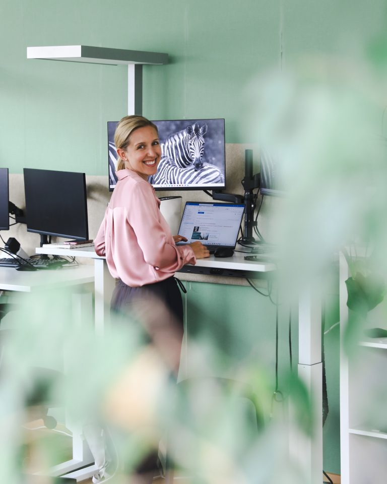 Blonde Frau steht an einem Schreibtisch und lächelt in die Kamera