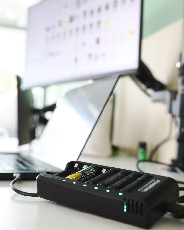 Ladegerät für Batterien vor einem Laptop und einem Bildschirm auf einem Schreibtisch